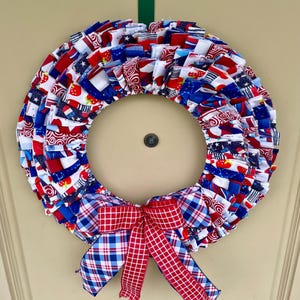May include: A red, white, and blue fabric wreath with a plaid bow. The wreath is made of various patriotic fabrics with stars, stripes, and strawberries.