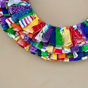 May include: A rainbow-colored fabric wreath with a variety of patterns and textures. The wreath is made of strips of fabric that are tied together to create a fringe effect.