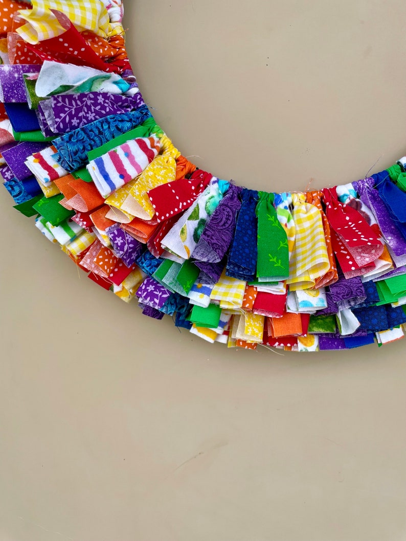 May include: A rainbow-colored fabric wreath with a variety of patterns and textures. The wreath is made of strips of fabric that are tied together to create a fringe effect.