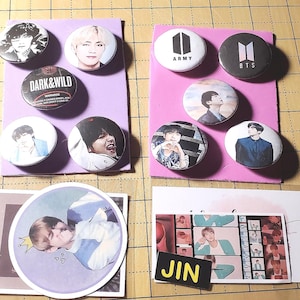 Può includere: Una collezione di otto spille rotonde con immagini di una band K-pop. Le spille sono su due carte rosa e bianche e due carte bianche. Le spille presentano immagini dei membri della band, il logo della band e le parole "Dark & Wild", "Army" e "BTS".