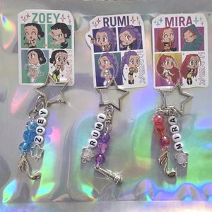 May include: Three keychains with star-shaped charms and beaded accents. Each keychain features a different name spelled out in white letter beads: Zoey, Rumi, and Mira. The keychains also include cartoon character stickers and musical note charms.
