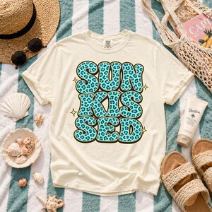 以下が含まれることがあります： 「SUN KISSED」の文字が大きくターコイズブルーのヒョウ柄でプリントされた、クリーム色のTシャツ。ストライプのタオル、麦わら帽子、サングラス、貝殻、サンダル、日焼け止めクリームと一緒に置かれています。ビーチをテーマにした夏のトップス。