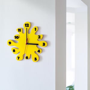 Könnte beinhalten: Eine moderne, gelbe Wanduhr mit einem einzigartigen, spritzartigen Design. Die Uhr hat schwarze Zahlen und Zeiger, mit einem roten Sekundenzeiger. Sie ist an einer weißen Wand montiert.