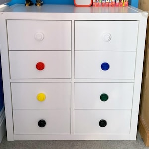 May include: White dresser with six drawers, each with a different colored knob. The knobs are red, blue, yellow, green, black, and white.