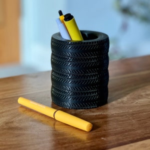 Op de afbeelding: Een zwarte, bandvormige potloodhouder met drie pennen erin. De pennen zijn geel en lichtblauw. De houder staat op een houten oppervlak.