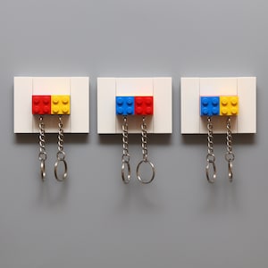 Pode incluir: Três porta-chaves retangulares brancos com um único chaveiro cada. Cada porta-chaves tem um par de blocos de Lego presos ao topo. Os blocos de Lego são vermelho e amarelo, azul e amarelo e azul e vermelho.
