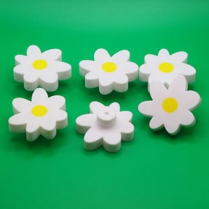 May include: Six white daisy-shaped cabinet knobs with yellow centers are arranged on a bright green background. One knob has a screw post on the back. The knobs are made of a smooth, solid material.