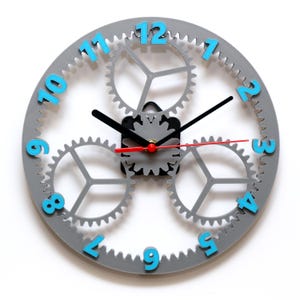 May include: Gray wall clock with three interlocking gears. The clock face has blue numbers and black hands. The clock is 12 inches in diameter.