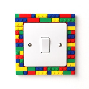 May include: A square light switch cover plate featuring a border constructed from small, interlocking plastic building bricks in primary colors including red, blue, yellow, and green. The central switch component is white with two metal screws.