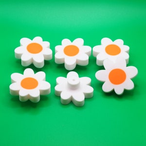 Op de afbeelding: Zes witte margrietvormige ladegrepen met oranje centra. De grepen zijn in een halve cirkel op een groene achtergrond gerangschikt.