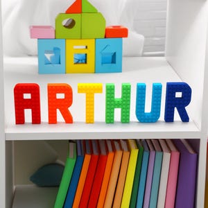 May include: A white shelf displays the name "ARTHUR" in colorful block letters. Above, a small house made of blocks in various colors sits. Below, a row of colorful books adds to the playful arrangement.