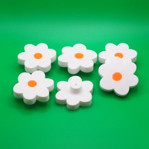Peut inclure: Six boutons en plastique blanc en forme de fleur avec des centres orange. Les boutons sont disposés en demi-cercle sur un fond vert.