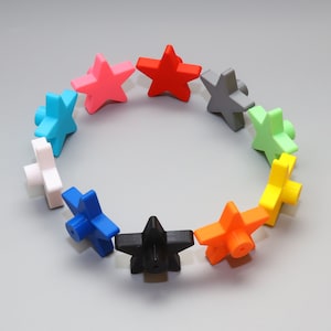 Może przedstawiać: Kolekcja kolorowych obiektów w kształcie gwiazd ułożonych w okrąg. Gwiazdy są w różnych kolorach, w tym czerwonym, różowym, niebieskim i żółtym. Każda gwiazda ma cylindryczną podstawę. Obiekty znajdują się na szarym tle.