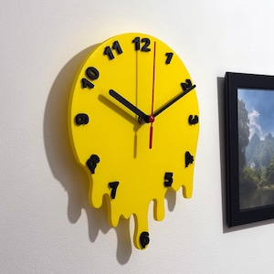 Pode incluir: Um relógio de parede amarelo, derretendo, com números e ponteiros pretos. O relógio tem um design único, gotejante, com a parte inferior parecendo derreter. O ponteiro dos segundos é vermelho. O relógio está montado em uma parede branca.