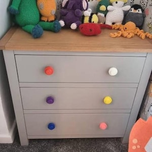 May include: A gray dresser with four drawers and colorful knobs. The knobs are orange, white, purple, yellow, blue, and pink. The dresser is decorated with crocheted stuffed animals.