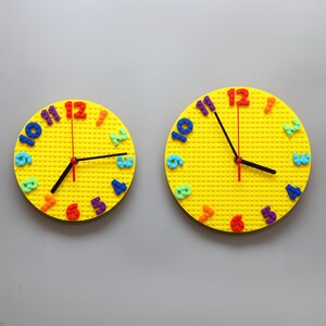 May include: Two yellow wall clocks with colorful numbers. The clocks have black hands and red second hands. The clocks are round and have a textured surface. The numbers are in various colors, including blue, green, orange, and purple.
