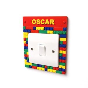May include: A white single light switch plate cover surrounded by a rectangular frame made of colorful red, blue, yellow, and green plastic building blocks. The top section features a red panel with the name OSCAR printed in yellow letters.