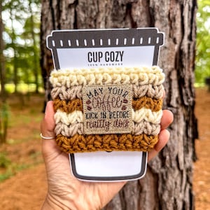 May include: A handmade cup cozy with the text "MAY YOUR COFFEE KICK IN BEFORE REALITY DOES." The cozy is made of tan, brown, and cream-colored yarn and is wrapped around a cardboard cup holder. The holder has the words "CUP COZY" printed on it.