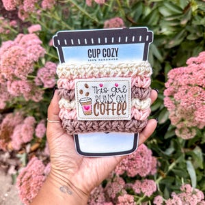 May include: A handmade cup cozy in shades of pink and brown, with the text "This girl runs on coffee." The cozy is designed to fit around a coffee cup. The background features pink flowers and green foliage.