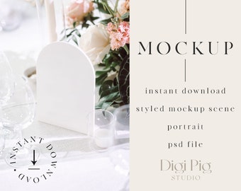 Peut inclure: Une maquette de carte de table blanche en forme d'arche sur une table blanche avec des fleurs roses et blanches, des verres transparents et des bougies. Le texte "MOCKUP" est affiché à droite de l'image avec les mots "téléchargement instantané", "scène de maquette stylisée", "portrait", "fichier psd", "Digi Pig STUDIO".