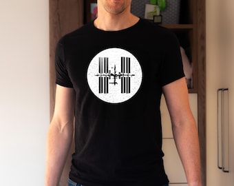 Op de afbeelding: Zwart T-shirt met een witte, verweerde cirkel met een zwart-witte afbeelding van het International Space Station. Het T-shirt heeft een ronde hals.