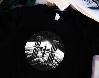 Op de afbeelding: Zwart t-shirt met een cirkelvormige zwart-wit afbeelding van het International Space Station (ISS) tegen een sterrenhemel en de aarde. Andere witte en groene t-shirts zijn zichtbaar op de achtergrond.