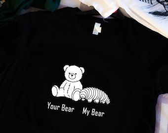 Puede incluir: Camiseta negra con un gráfico blanco de un oso de peluche y un gusano. El texto "Your Bear" y "My Bear" está impreso debajo del gráfico.