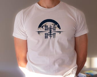 Op de afbeelding: Een wit T-shirt met een afbeelding van een ruimtestation dat wordt weerspiegeld in een watermassa, met een halve maan en sterren op de achtergrond.
