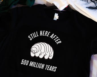 Puede incluir: Camiseta negra con un gráfico blanco de un tardígrado, un animal microscópico, con el texto "Still Here After 500 Million Years".
