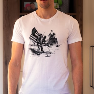 May include: A white t-shirt with a black and white illustration of an astronaut planting an American flag on the moon. The astronaut is standing next to a lunar module.
