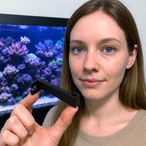 May include: A black, L-shaped bracket with a hook on one end is held up. The bracket appears to be made of a solid material. The background features an aquarium with colorful coral and plants.