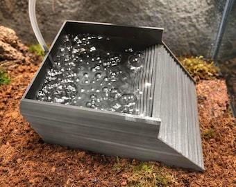 May include: A gray, angular water feature with a rippled side and a basin filled with bubbling water. The water feature is set on a bed of brown and green substrate, suggesting a terrarium or similar environment.