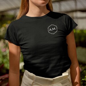 Könnte beinhalten: Schwarzes T-Shirt mit Rundhalsausschnitt und weißem geometrischen Design sowie den Buchstaben "A.M." auf der Brust. Das Shirt besteht aus einem weichen Material und hat kurze Ärmel. Das Model trägt einen weißen Rock.
