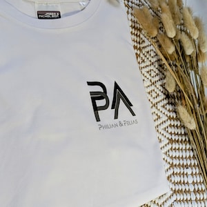K&ouml;nnte beinhalten: Ein wei&szlig;es T-Shirt mit dem schwarzen Text "PA" und dem Markennamen "PHILIAN & FELIAS" darunter. Das T-Shirt hat einen Rundhalsausschnitt und ein Etikett mit der Aufschrift "JAMES & NICHOLSON".