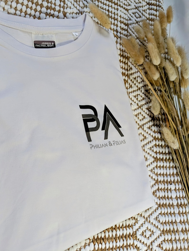 K&ouml;nnte beinhalten: Ein wei&szlig;es T-Shirt mit dem schwarzen Text "PA" und dem Markennamen "PHILIAN & FELIAS" darunter. Das T-Shirt hat einen Rundhalsausschnitt und ein Etikett mit der Aufschrift "JAMES & NICHOLSON".