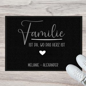 Könnte beinhalten: Schwarze Fußmatte mit dem Wort "Familie" in Schreibschrift, darunter der Satz "IST DA, WO DAS HERZ IST". Ein weißes Herz und die Namen "Melanie + Alexander" sind ebenfalls abgebildet. Weiße Turnschuhe sind in der unteren rechten Ecke zu sehen.