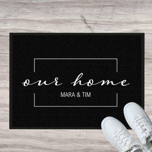 Könnte beinhalten: Schwarze Willkommensmatte mit weißem Text, der "our home" und "Mara & Tim" in einem weißen Rechteck lautet.