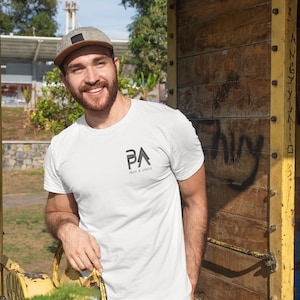 K&ouml;nnte beinhalten: Wei&szlig;es T-Shirt mit dem Text "PA" und "MAN & DADS" in Schwarz gedruckt. Die Person auf dem Bild tr&auml;gt eine grau-braune Baseballkappe. Der Hintergrund umfasst eine gelbe Metallstruktur und eine Holzwand.