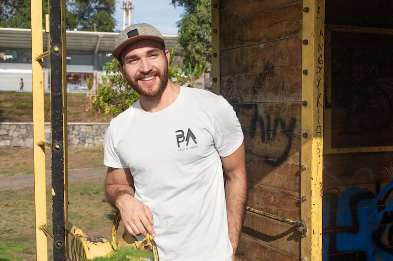 K&ouml;nnte beinhalten: Wei&szlig;es T-Shirt mit dem Text "PA" und "MAN & DADS" in Schwarz gedruckt. Die Person auf dem Bild tr&auml;gt eine grau-braune Baseballkappe. Der Hintergrund umfasst eine gelbe Metallstruktur und eine Holzwand.