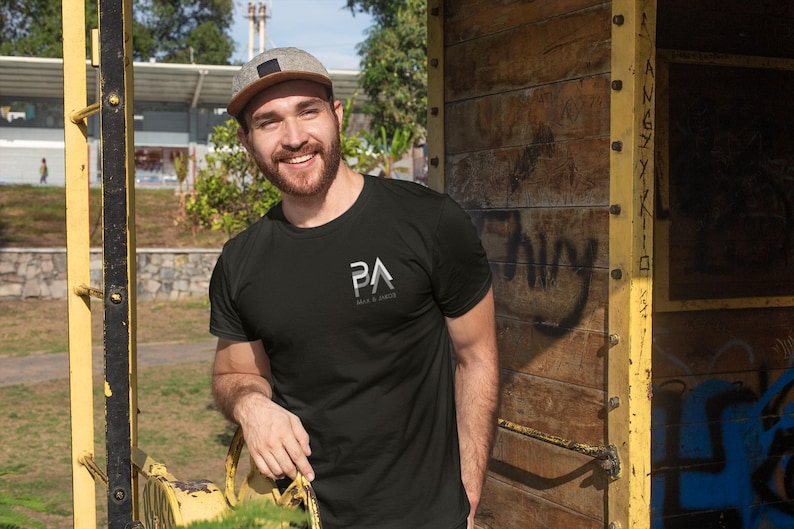 K&ouml;nnte beinhalten: Ein schwarzes T-Shirt mit einem kleinen wei&szlig;en Logo auf der Brust. Die Person auf dem Bild tr&auml;gt eine grau-braune Baseballkappe. Der Hintergrund umfasst eine Holzkonstruktion und eine gelbe Metallkonstruktion.