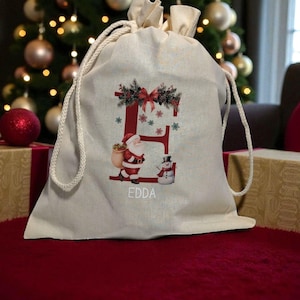 Könnte beinhalten: Beiger Weihnachtsgeschenkbeutel mit Kordelzug und festlichem Design. Der Beutel zeigt einen großen roten Buchstaben "E", verziert mit Stechpalme und einer Schleife. Der Weihnachtsmann und ein Schneemann sind ebenfalls abgebildet. Der Name "EDDA" ist darunter gedruckt.