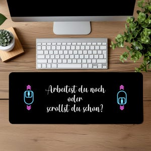 Könnte beinhalten: Schwarze Mauspad mit weißem Text, der "Arbeitest du noch oder scrollst du schon?" lautet, mit einem blauen und pinkfarbenen Maussymbol auf jeder Seite.