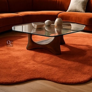 Pode incluir: Um tapete laranja, com formato org&acirc;nico, est&aacute; sobre um piso de madeira, sob uma mesa de centro com tampo de vidro e base de madeira. Um sof&aacute; laranja curvo est&aacute; ao fundo. H&aacute; objetos decorativos na mesa.