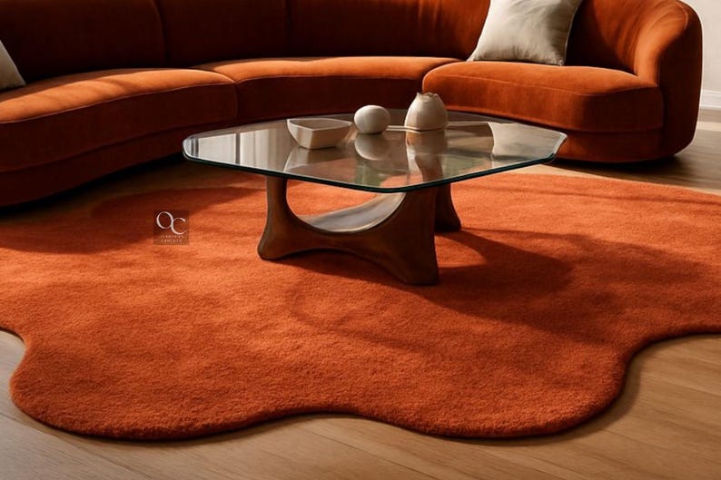 Pode incluir: Um tapete laranja, com formato org&acirc;nico, est&aacute; sobre um piso de madeira, sob uma mesa de centro com tampo de vidro e base de madeira. Um sof&aacute; laranja curvo est&aacute; ao fundo. H&aacute; objetos decorativos na mesa.