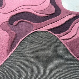 May include: A plush, abstract rug in shades of pink and purple. The rug features a layered design with varying textures and depths, creating a three-dimensional effect. The underside of the rug is visible, showing a dark gray backing.