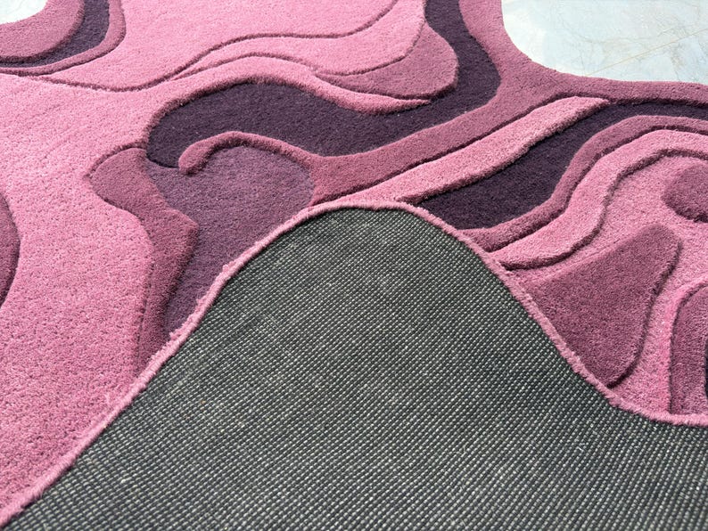 May include: A plush, abstract rug in shades of pink and purple. The rug features a layered design with varying textures and depths, creating a three-dimensional effect. The underside of the rug is visible, showing a dark gray backing.