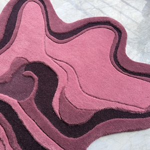 May include: A plush, abstract rug in shades of pink and purple. The rug features a layered design with varying textures and tones, creating a unique, organic shape. The rug's design resembles a topographical map.