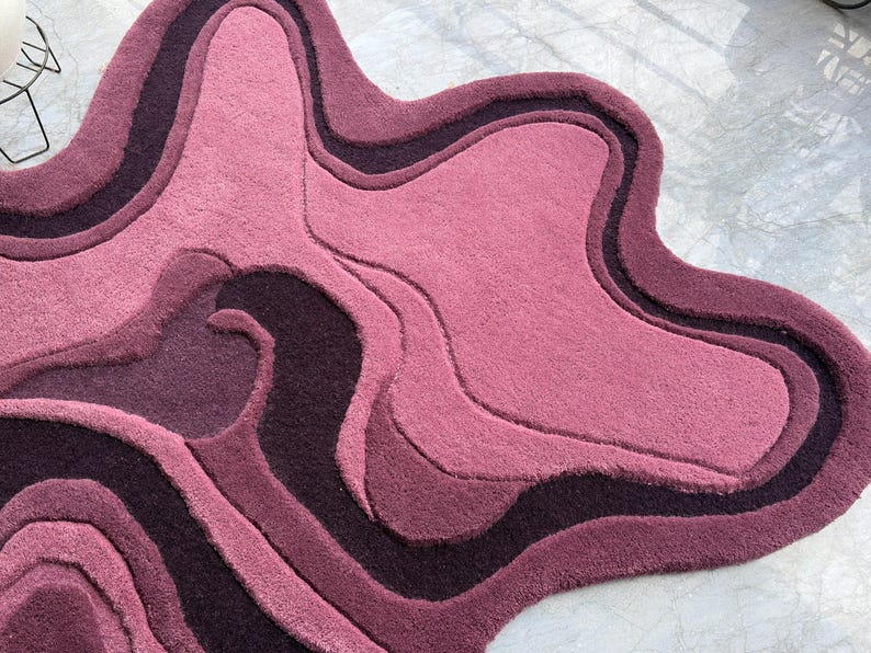 May include: A plush, abstract rug in shades of pink and purple. The rug features a layered design with varying textures and tones, creating a unique, organic shape. The rug's design resembles a topographical map.
