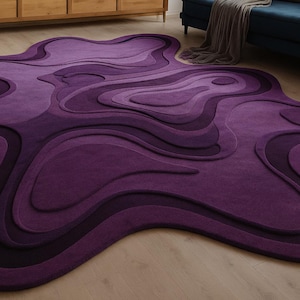 May include: A large, abstract area rug in shades of purple. The rug features a layered, organic design with varying depths and textures. The color palette ranges from deep violet to lighter lavender hues, creating a visually dynamic piece for a living space.