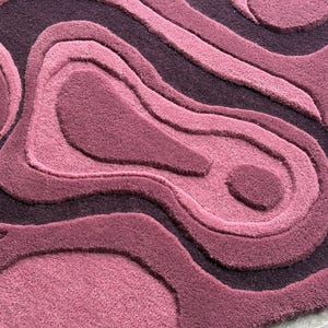 May include: A plush, abstract rug in shades of purple and mauve. The rug features a layered, topographic design with varying pile heights, creating a textured, three-dimensional effect. The rug's design resembles contour lines, adding visual interest to any space.